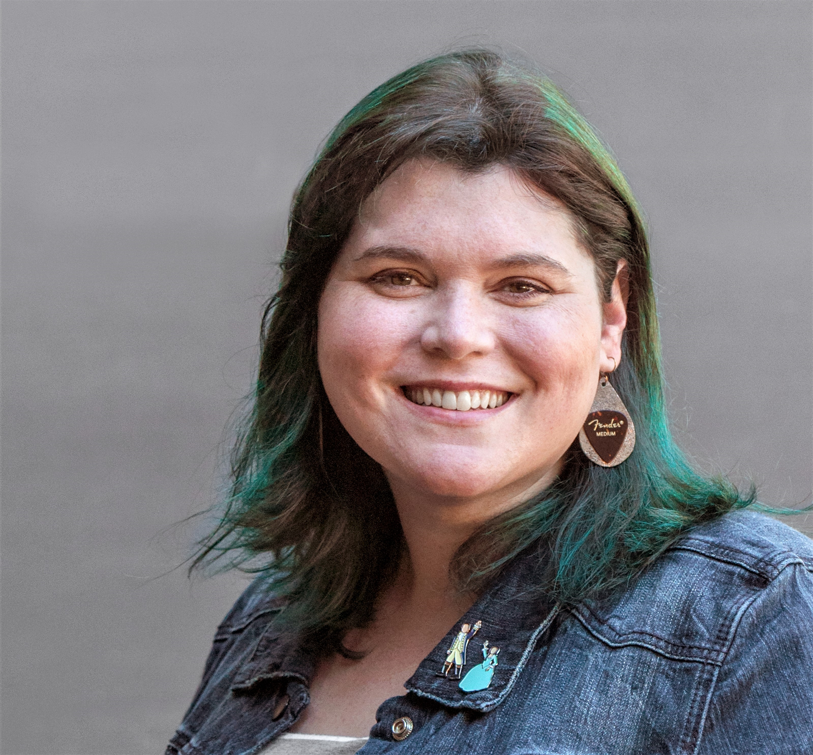 Headshot of Amber Bracegirdle wearing a denim jacket, earrings with guitar pics on them, with teal highlighted hair.