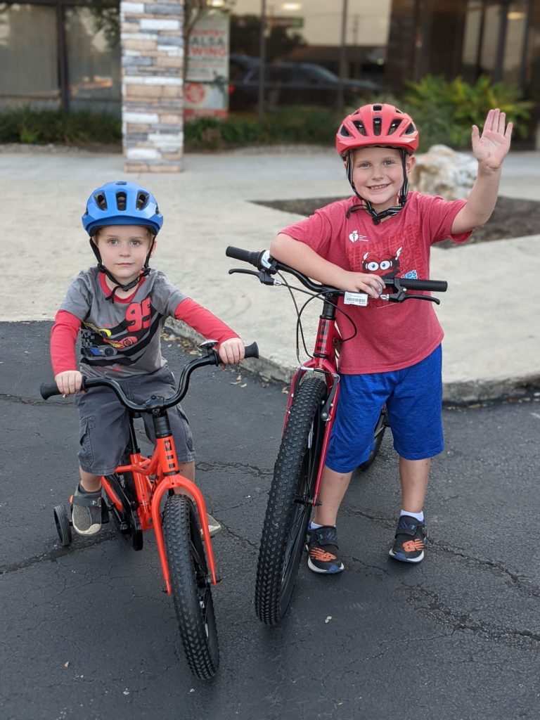 Two smiling boys look at the camera. They are both in bike helmets and sitting or standing next to bicycles.