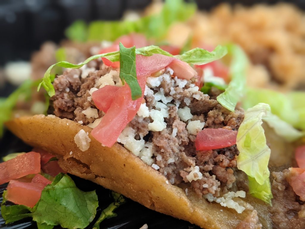 A Tex-Mex Puffy Taco photographed closely. There are tomatoes and lettuce as well as picadillo (ground beef and potatoes in a savory sauce).